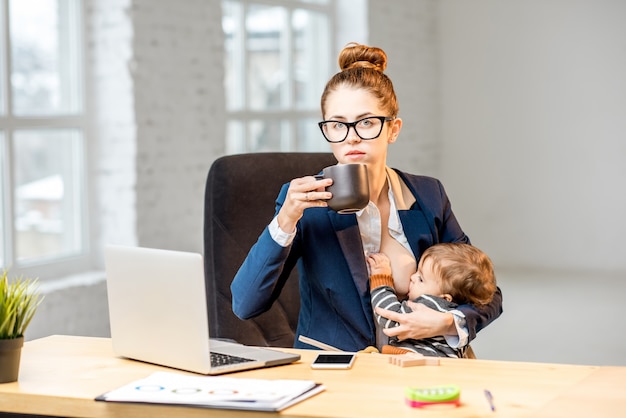 Foto jonge multitasking zakenvrouw die haar zoontje borstvoeding geeft terwijl ze een pauze koffie drinkt op kantoor