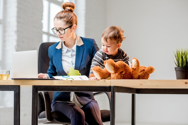 Jonge multitasking zakenvrouw gekleed in het pak werkend met laptop en documenten zittend met haar zoontje op kantoor