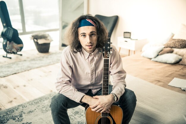 Jonge muzikant rusten. aangename professionele gitarist in licht shirt en bandana terwijl hij zijn persoonlijke instrument vasthoudt