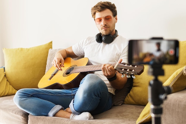 Jonge muzikantblogger met een bril die op de bank voor een camera zit, speelt terloops akoestische gitaar in een helder verlichte kamer