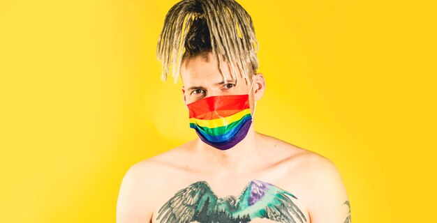 Jonge naakte man met tatoeage op borst in Lgbt-masker camera kijken op gele achtergrond knappe jongen met dreadlocks in beschermende regenboog masker poseren in studio