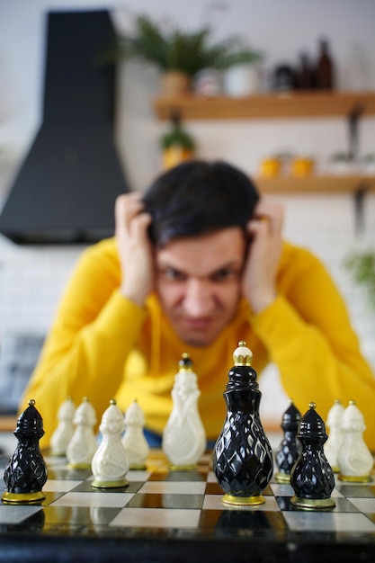 Jonge nadenkende man aan het schaken op de keukentafel Peinzende man speelt in logisch bordspel met zichzelf in de keuken