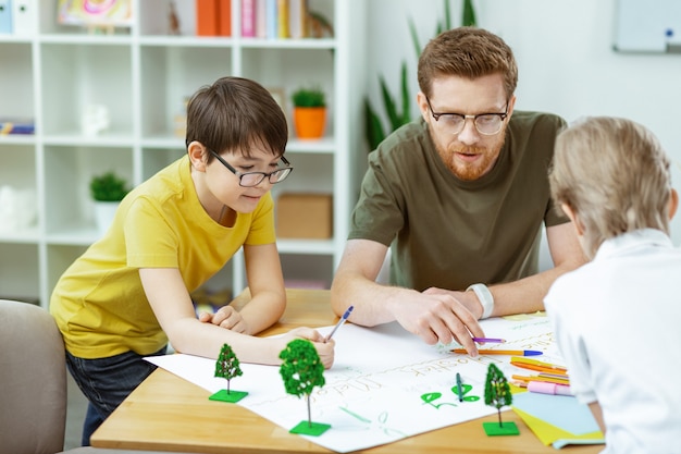 Jonge natuurliefhebbers. Attente geïnspireerde bebaarde leraar die zijn leerlingen helpt met handgemaakte poster terwijl ze allemaal samen zitten sitting