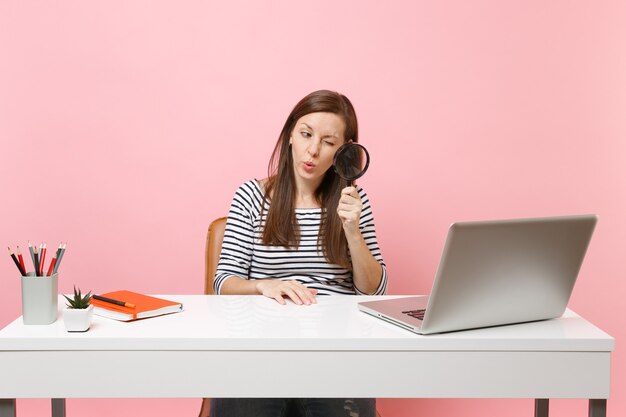 Jonge nieuwsgierige vrouw die door vergrootglas op pc-laptop kijkt terwijl ze aan project op kantoor werkt geïsoleerd op pastelroze achtergrond. prestatie zakelijke carrière concept. ruimte kopiëren.