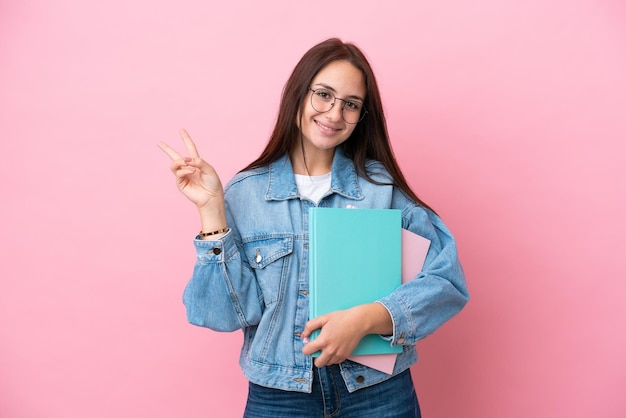 Jonge Oekraïense studentenvrouw die op roze achtergrond glimlacht en overwinningsteken toont