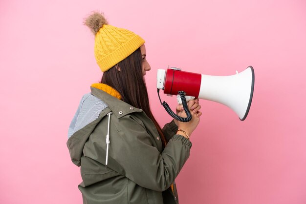 Jonge Oekraïense vrouw die winterjas draagt die op roze achtergrond wordt geïsoleerd en door een megafoon schreeuwt