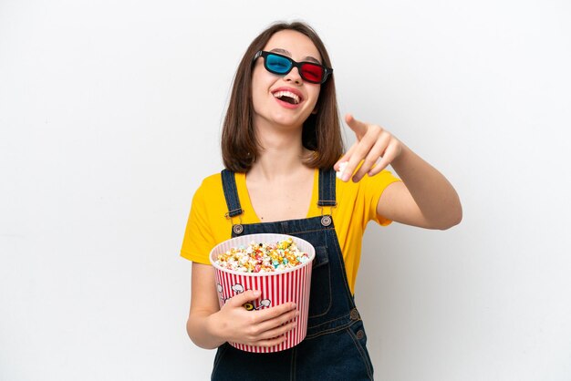 Jonge Oekraïense vrouw geïsoleerd op een witte achtergrond met een 3D-bril en een grote emmer popcorns vasthoudend terwijl ze naar voren wijst