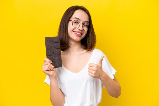 Jonge Oekraïense vrouw geïsoleerd op gele achtergrond neemt een chocoladetablet en met duim omhoog