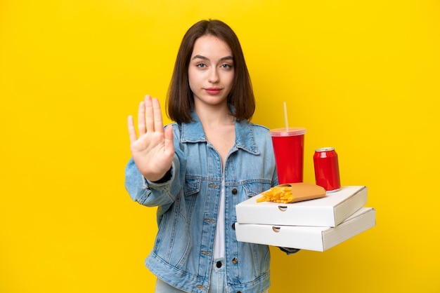 Jonge Oekraïense vrouw met fastfood geïsoleerd op een gele achtergrond die stopgebaar maakt