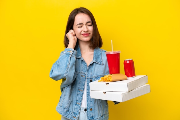 Jonge Oekraïense vrouw met fastfood geïsoleerd op gele achtergrond gefrustreerd en oren bedekkend