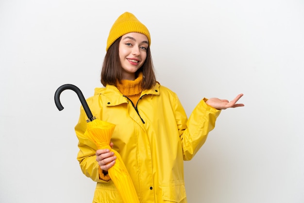 Jonge Oekraïense vrouw met regenbestendige jas en paraplu geïsoleerd op een witte achtergrond die de handen naar de zijkant uitstrekt om uit te nodigen om te komen