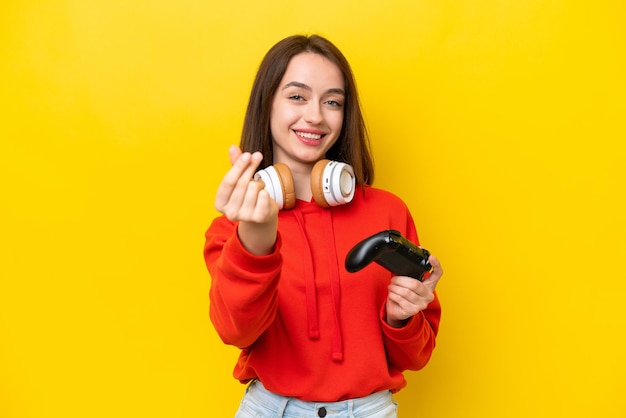 Jonge Oekraïense vrouw speelt met een videogamecontroller geïsoleerd op een gele achtergrond die geld gebaar maakt