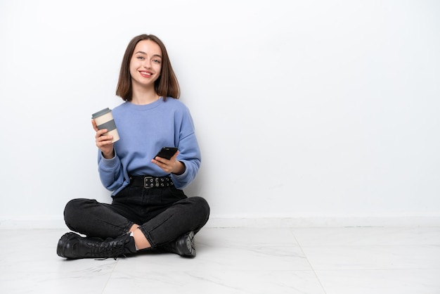 Jonge Oekraïense vrouw zittend op de vloer geïsoleerd op een witte achtergrond met koffie om mee te nemen en een mobiel