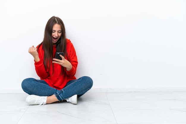 Jonge Oekraïense vrouw zittend op de vloer geïsoleerd op een witte achtergrond verrast en het verzenden van een bericht