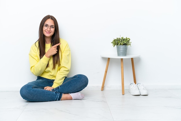 Jonge Oekraïense vrouw zittend op de vloer van haar huis geïsoleerd op een witte muur wijzend naar de zijkant om een product te presenteren