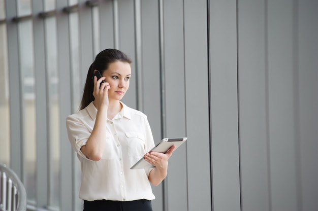 Jonge onderneemster die op mobiele telefoon spreekt terwijl status door venster in bureau. Mooi jong vrouwelijk model in een helder bureau.