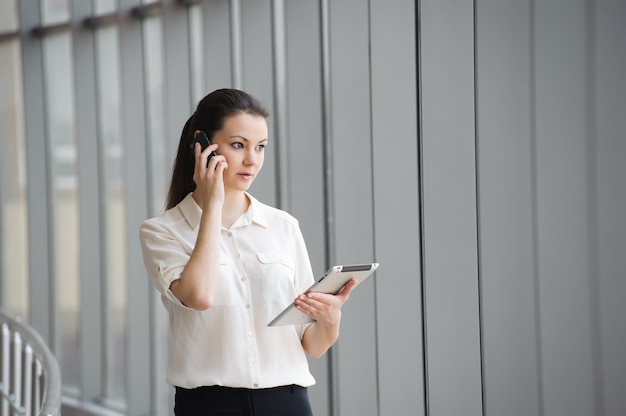 Jonge onderneemster die op mobiele telefoon spreekt terwijl status door venster in bureau. Mooie jonge vrouwelijke model in office.