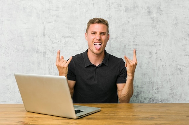 Jonge ondernemer die met zijn laptop aan een bureau werkt dat rotsgebaar met vingers toont