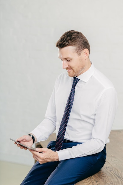 Jonge ongeschoren intelligente man draagt formele kleding gebruikt digitale tablet voor werk gericht op scherm zit aan bureau geïsoleerd op witte achtergrond programmeur ontwikkelt applicatiesoftware