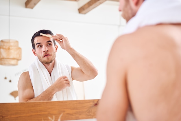 Jonge ongeschoren man die zijn haar kamt terwijl hij met een witte handdoek om zijn nek bij de spiegel in de badkamer staat