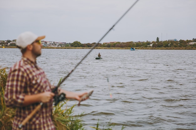 Jonge ongeschoren man in geruit hemd, pet, zonnebril uitgetrokken hengel en houdt gevangen vis aan de oever van het meer in de buurt van riet op de achtergrond van de boot. Lifestyle, recreatie, vrijetijdsconcept voor vissers