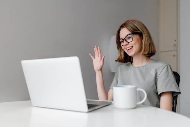 Jonge ontspannen blanke vrouw die lacht zwaaien met laptop in het appartement met kopje op tafel