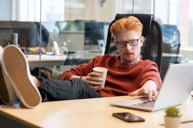 Jonge ontspannen kantoormedewerker in vrijetijdskleding met een glas koffie tijdens de pauze terwijl hij in een fauteuil zit met zijn benen op tafel voor de laptop