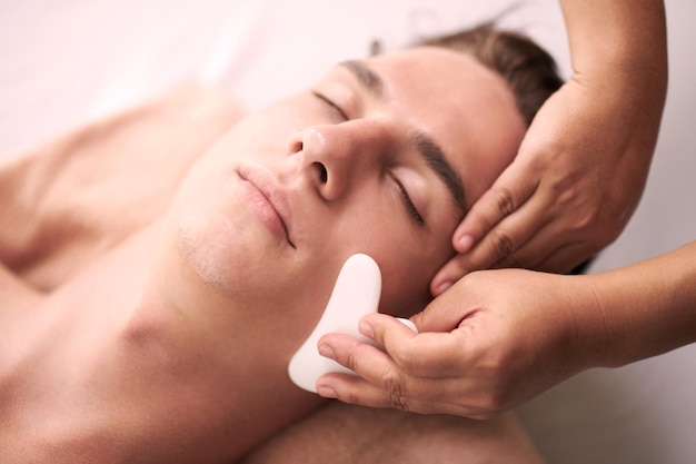 Jonge ontspannen mannelijke klant van een spa salon krijgt een luxe gua sha gezichtsmassage bwing gegeven aan hem door een professionele vrouwelijke masseuse
