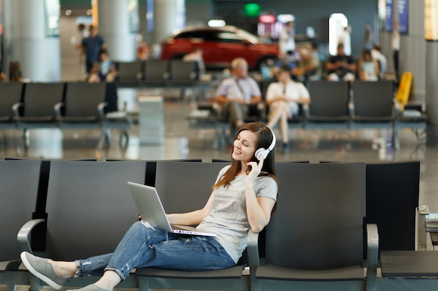Jonge ontspannen reizigerstoeristenvrouw met koptelefoon die muziek luistert die op laptop werkt, wacht in de lobby op de internationale luchthaven