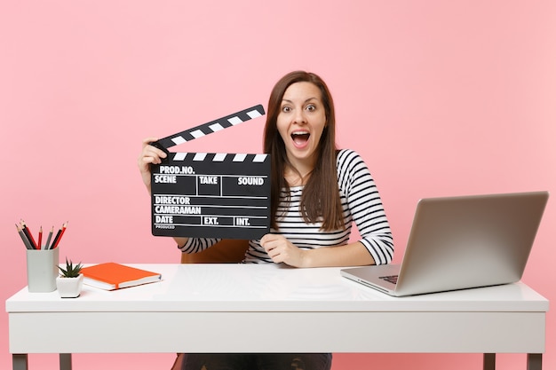 Jonge opgewonden vrouw houdt klassieke zwarte film vast en maakt filmklapper die aan een project werkt terwijl ze op kantoor zit met een laptop