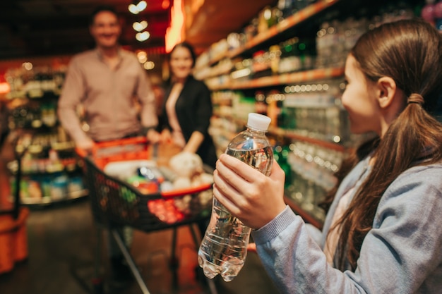 Jonge ouders en dochter in de supermarkt. Het meisje houdt waterfles in handen en bekijkt ouders. Ze staan achter en dragen een boodschappenwagentje.