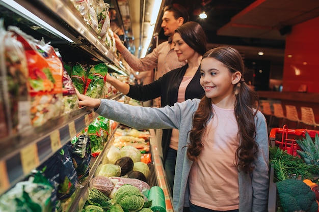 Jonge ouders en dochter in de supermarkt. Ze staan voor de slaplank en pakken het op. Mensen glimlachen. Gelukkige familie samen.