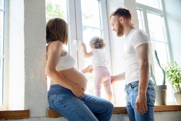 Jonge ouders man en zwangere vrouw spelen met dochtertje thuis en kijken uit het raam Concept van zwangerschap familie liefdesrelatie ouderschap en kindertijd