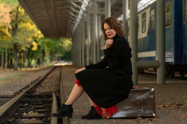 Jonge ouderwetse vrouw in zwarte jas zit op vintage koffer en wacht op trein Vrouw van middelbare leeftijd op treinstation