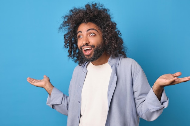 Jonge overgelukkige Arabische man spreidt handen naar de zijkant staat op blauwe achtergrond