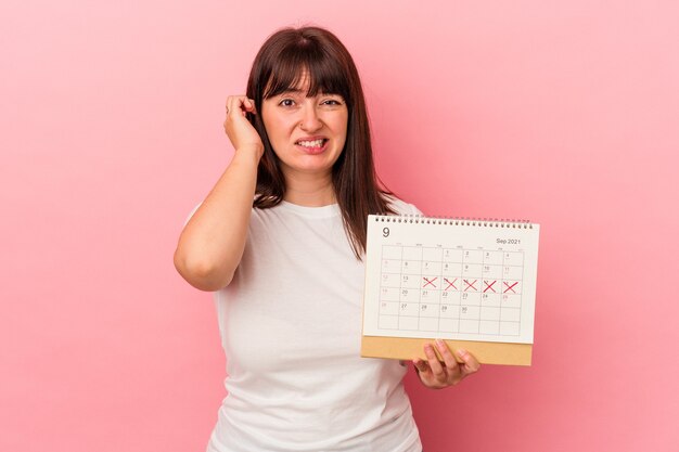 Jonge overgewicht blanke vrouw met kalender geïsoleerd op roze achtergrond die oren bedekt met handen.