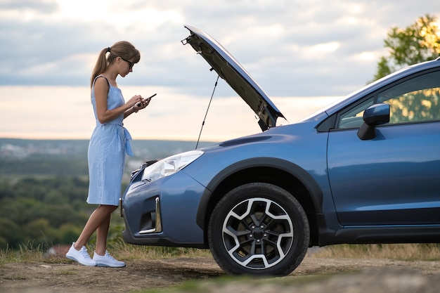 Jonge overstuur vrouw bestuurder praten op mobiele telefoon in de buurt van een kapotte auto met open motorkap inspecteren motor problemen met haar voertuig.