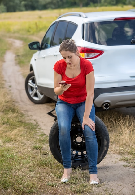 Foto jonge overstuur vrouw die op een reservewiel naast een kapotte auto op het veld zit en om hulp roept