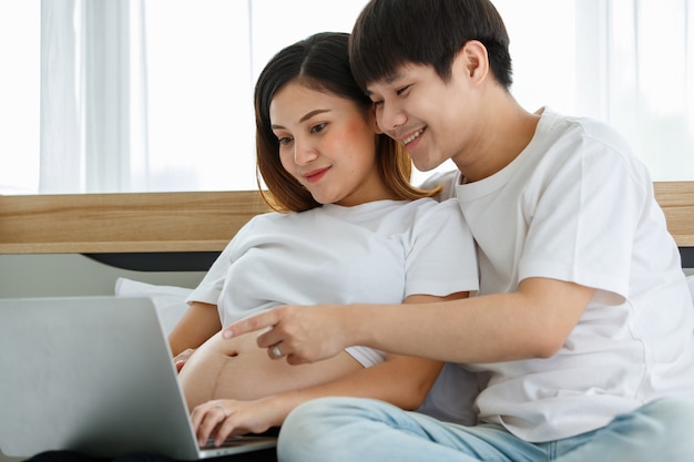 Jonge paar Aziatische man en zwangere vrouw zitten ontspannen op een bed met kijken naar een laptop op de schoot van een dame. Man sloeg een arm om zijn vrouw en wees naar een computerscherm. Familieconcept.