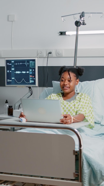 Jonge patiënt die video-oproep op laptop gebruikt voor communicatie op afstand terwijl hij in het ziekenhuisbed zit. Vrouw praat met vrienden op online videoconferentieconferentie over gezondheidszorg.