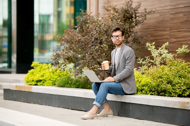 Jonge peinzende auteur in smart casual met een drankje terwijl hij nieuwe ideeën bedenkt voor een nieuw boek in een stedelijke omgeving