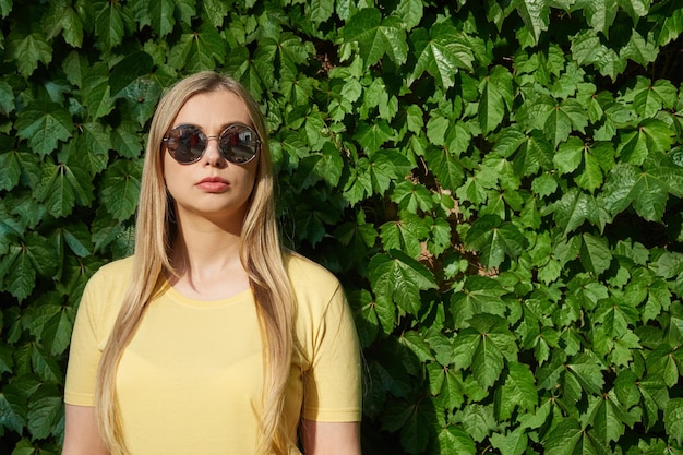 Jonge peinzende vrouw in een gele t-shirt in zonnebril staat tegen een achtergrond van groene natuurlijke fo