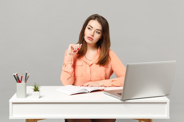 Jonge peinzende vrouw in pastel casual kleding die notities schrijft, zit en werkt aan een wit bureau met pc-laptop geïsoleerd op een grijze achtergrond. Prestatie zakelijke carrière levensstijl concept. Bespotten kopie ruimte.