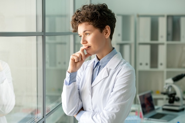 Jonge peinzende vrouwelijke wetenschapper of laboratoriummedewerker in whitecoat staande door glazen wand tegen haar werkplek met laptop en microscoop