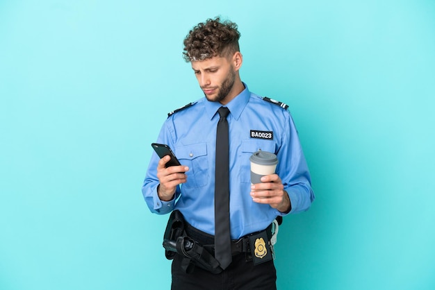 Jonge politie blonde man isoleerde wit op blauwe achtergrond met koffie om mee te nemen en een mobiel