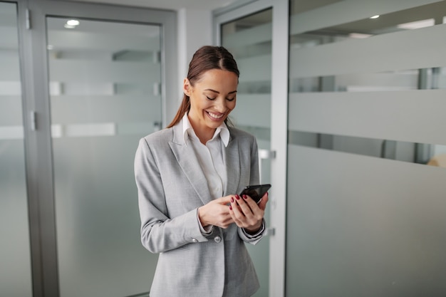 Jonge positieve blanke zakenvrouw in pak staande binnenkant van corporate firma en met behulp van slimme telefoon voor het lezen van een e-mail.