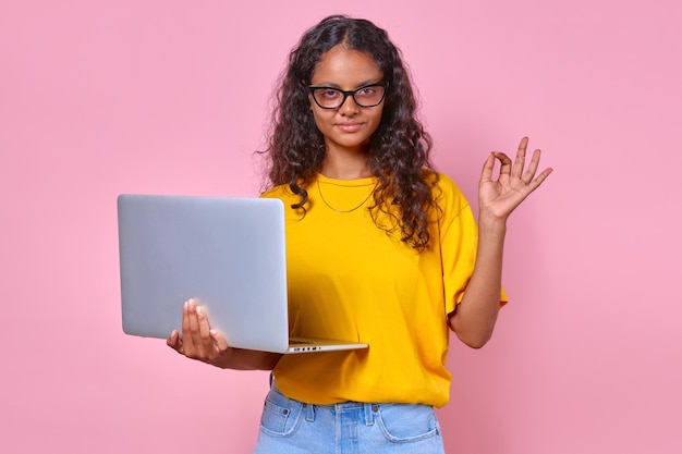 Jonge positieve Indiase tienervrouw met laptop in de hand maakt een ok gebaar