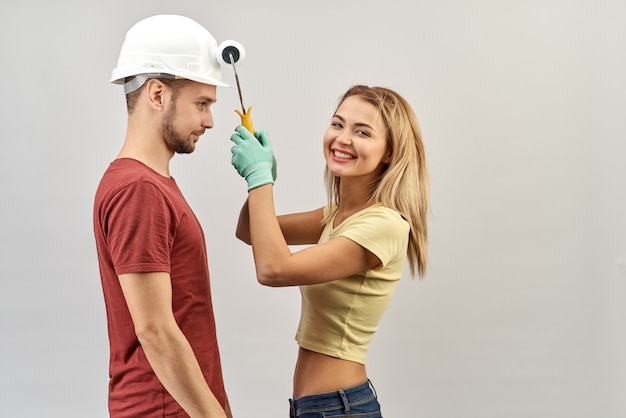 Jonge positieve paar gek rond tijdens het repareren en schilderen van de muren in het appartement. Een man in een witte helm en het meisje in handschoenen met verfroller in de hand