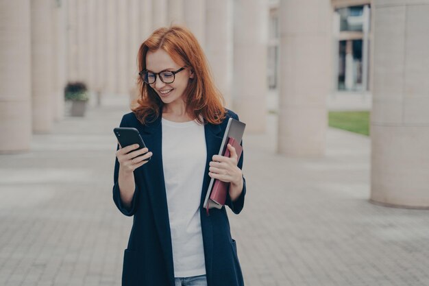 Jonge positieve roodharige zakenvrouw in elegante outfit met mobiele telefoon die buiten staat