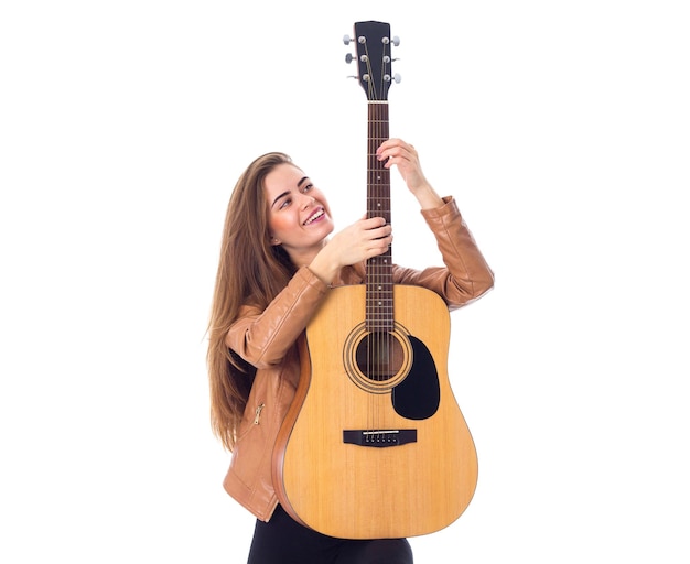 Jonge positieve vrouw die in bruin jasje een gitaar op witte achtergrond in studio houdt